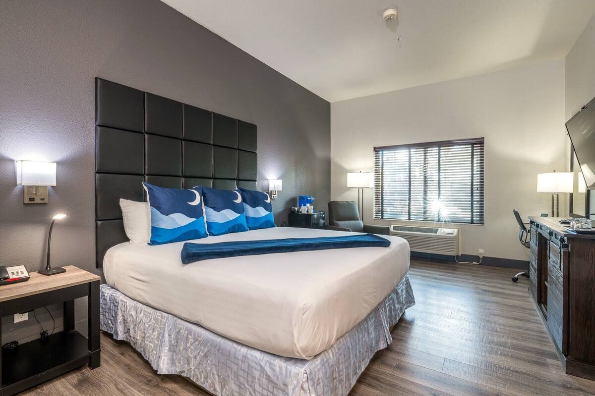 A hotel room with a large bed, a desk, a chair, a desk lamp, a nightstand, a wall-mounted lamp, a window with blinds, and a ceiling light.