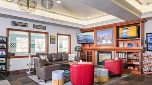 A hotel lobby with a leather sofa, red ottoman, and a fireplace. There are signs for 'HAILEY' and 'IDAHO' on the wall, and a television mounted above the fireplace. - Wood River Inn & Suites (Hailey)
