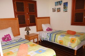 A hotel room with two single beds, wooden headboards, a small wooden nightstand, a lamp, and a wall with tropical-themed pictures.