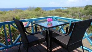A glass table with two wicker chairs on a deck with a blue railing, overlooking a coastal landscape. - West Hill Bungalows (Sabana Westpunt)