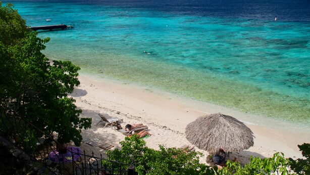 A beach with clear turquoise water, a thatched umbrella, and a dock. - West Hill Bungalows (Sabana Westpunt)