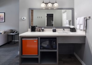A modern bathroom with a large mirror, a sink, and a compact microwave. There is a small table with a phone and a coffee maker, and a basket with toiletries.