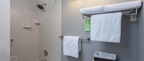 A modern bathroom with a bathtub, shower, and toilet. There are white towels on a rack, a green sign, and a basket on the toilet.