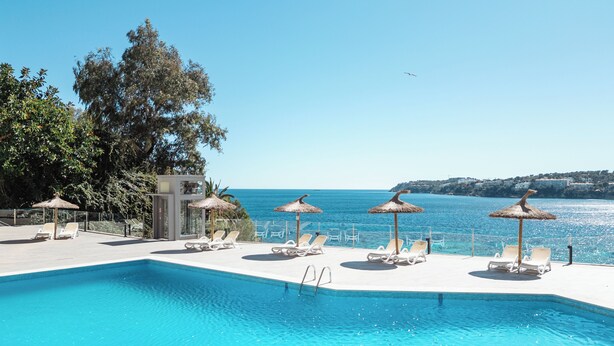 A poolside area with lounge chairs and umbrellas, a clear view of the sea, and a building with a glass facade.