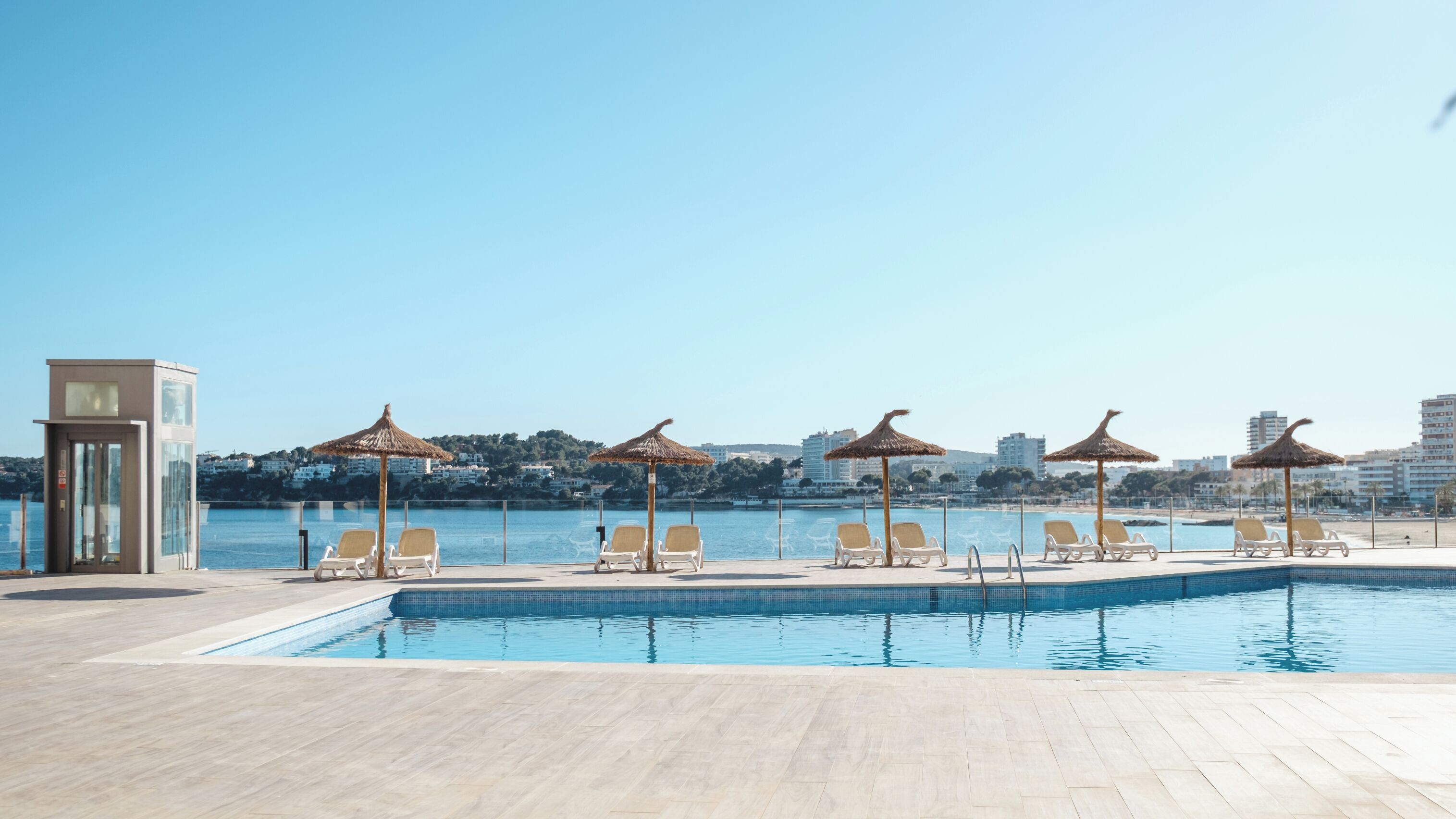 Una piscina con sillas de descanso y sombrillas, una torre de salvavidas y un paisaje urbano al fondo.