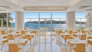 A spacious dining area with tables and chairs, large windows with a view of the sea and hills.