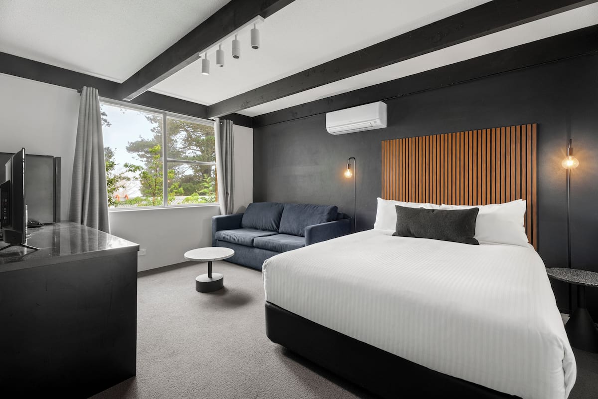A modern hotel room with a large bed, a desk, a sofa, and a view of the outdoors.