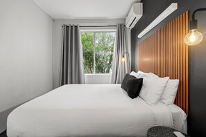 A modern hotel room with a large bed, a wooden headboard, a window with curtains, and a wall-mounted light fixture. - Motel Echo Point (Katoomba)
