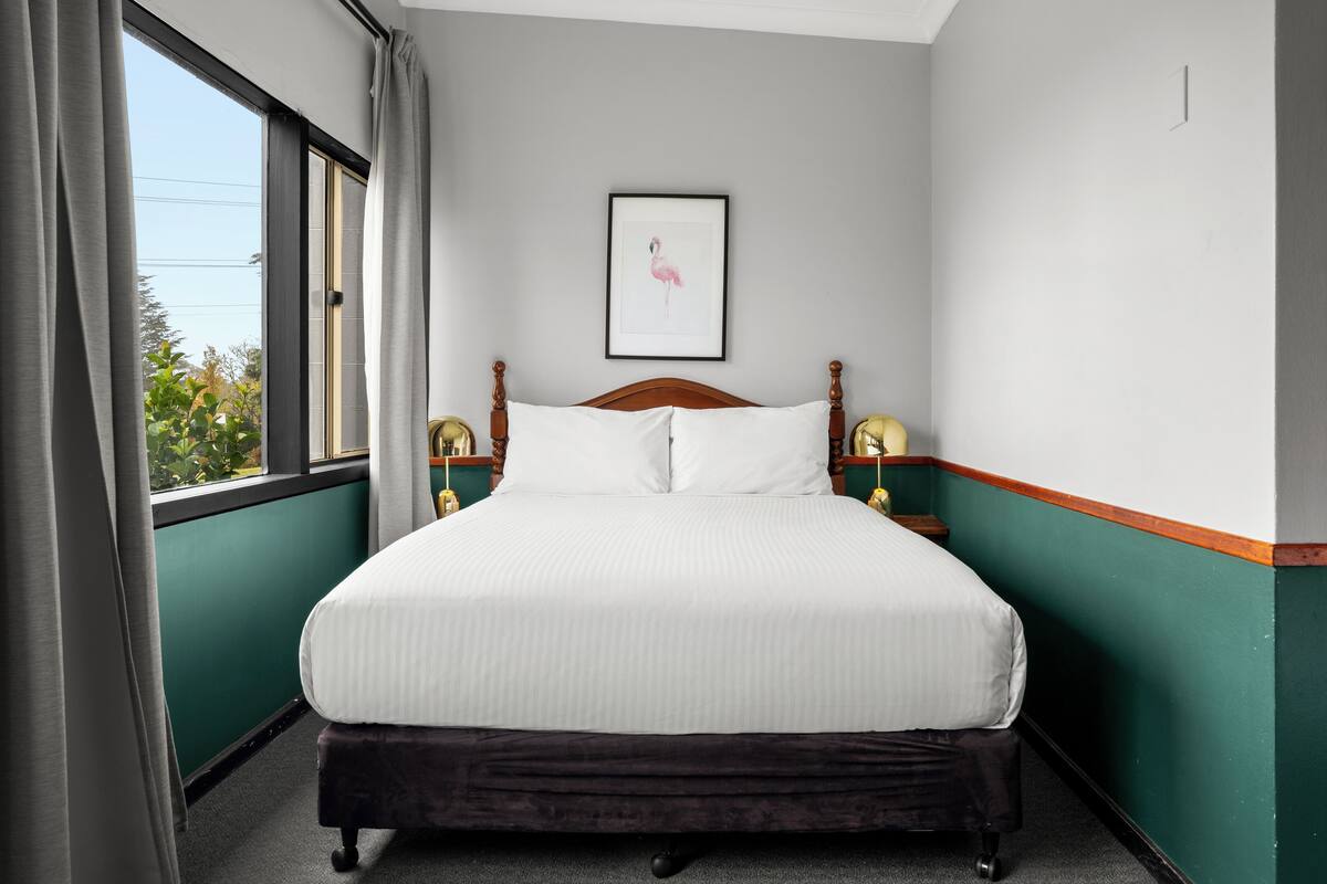 A neatly made bed with a wooden headboard, a framed picture on the wall, and a window with curtains.