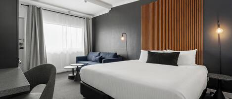 A modern hotel room with a large bed, a desk, a chair, and a sofa.