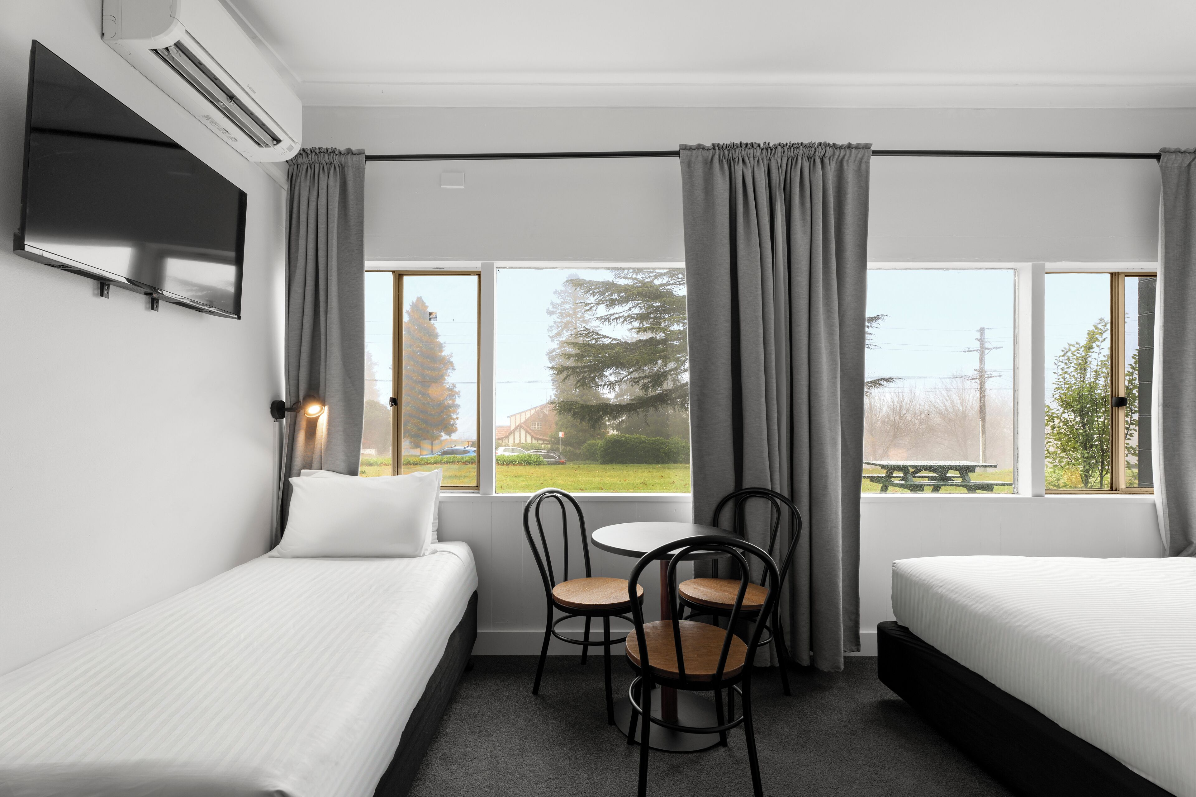 A hotel room with two single beds, a small table with chairs, a TV, and large windows with curtains.
