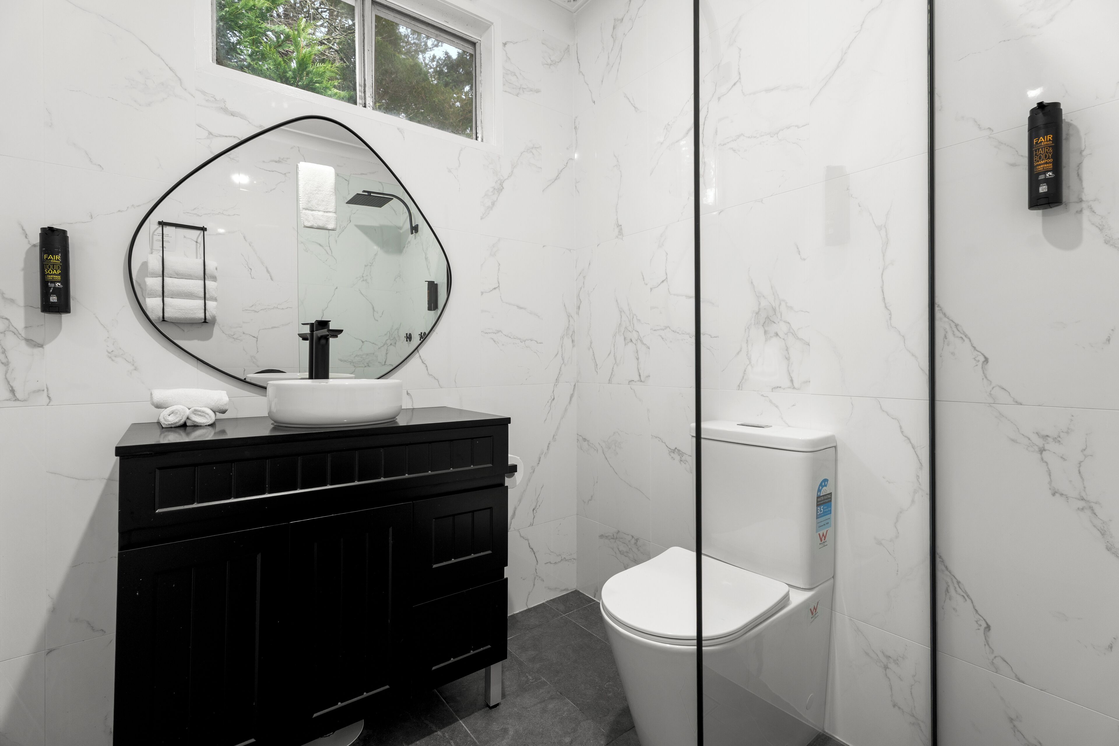 A modern bathroom with a black vanity, a round mirror, a white toilet, and a glass shower enclosure.