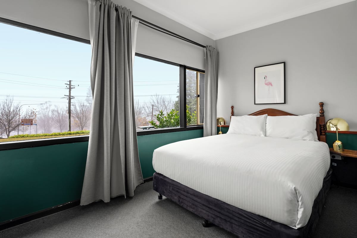 A hotel room with a large bed, a window with curtains, a framed picture on the wall, and a nightstand with a lamp.
