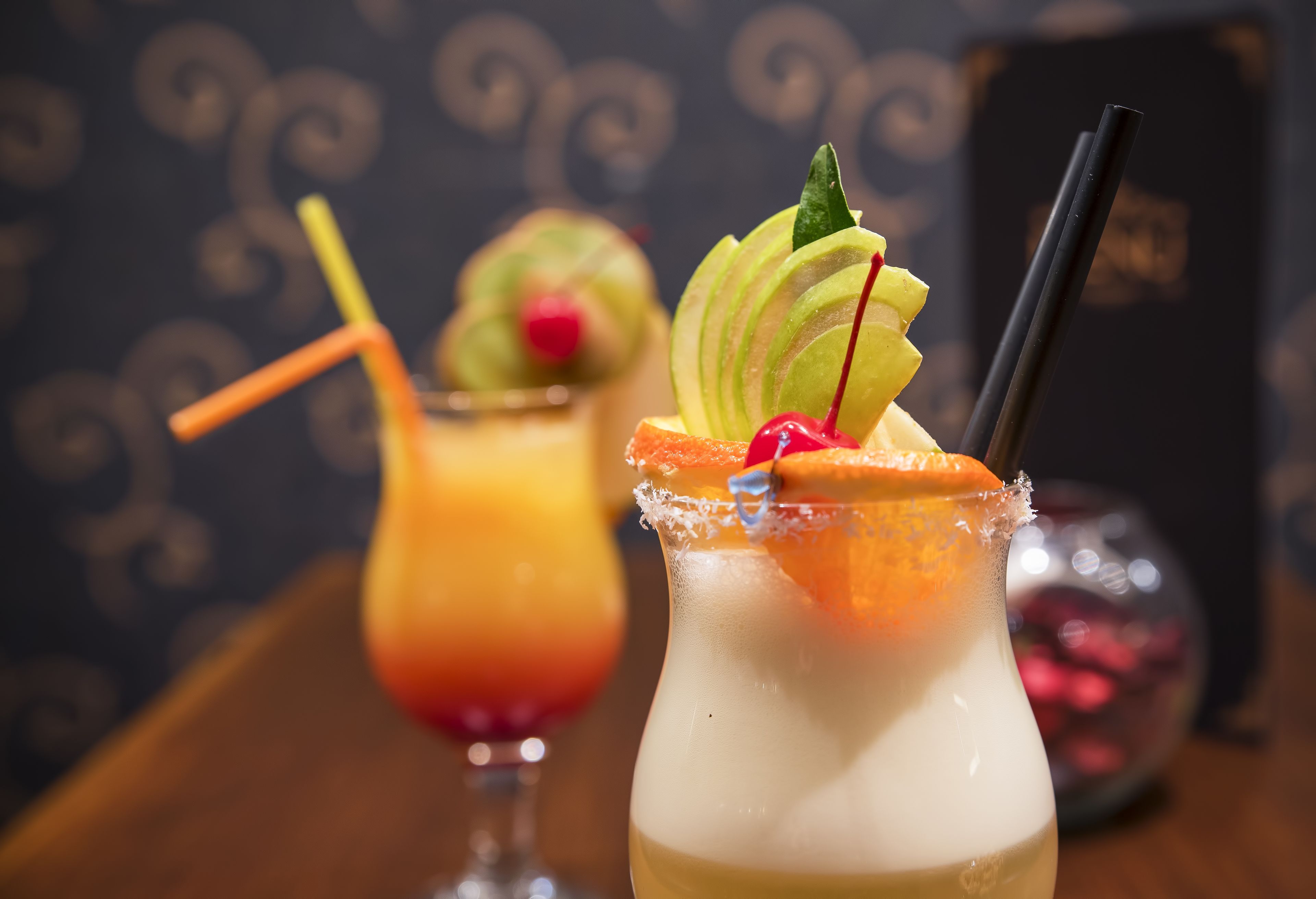 Two cocktails with garnishes and straws, a decorative glass, and a blurred background with a patterned wall.