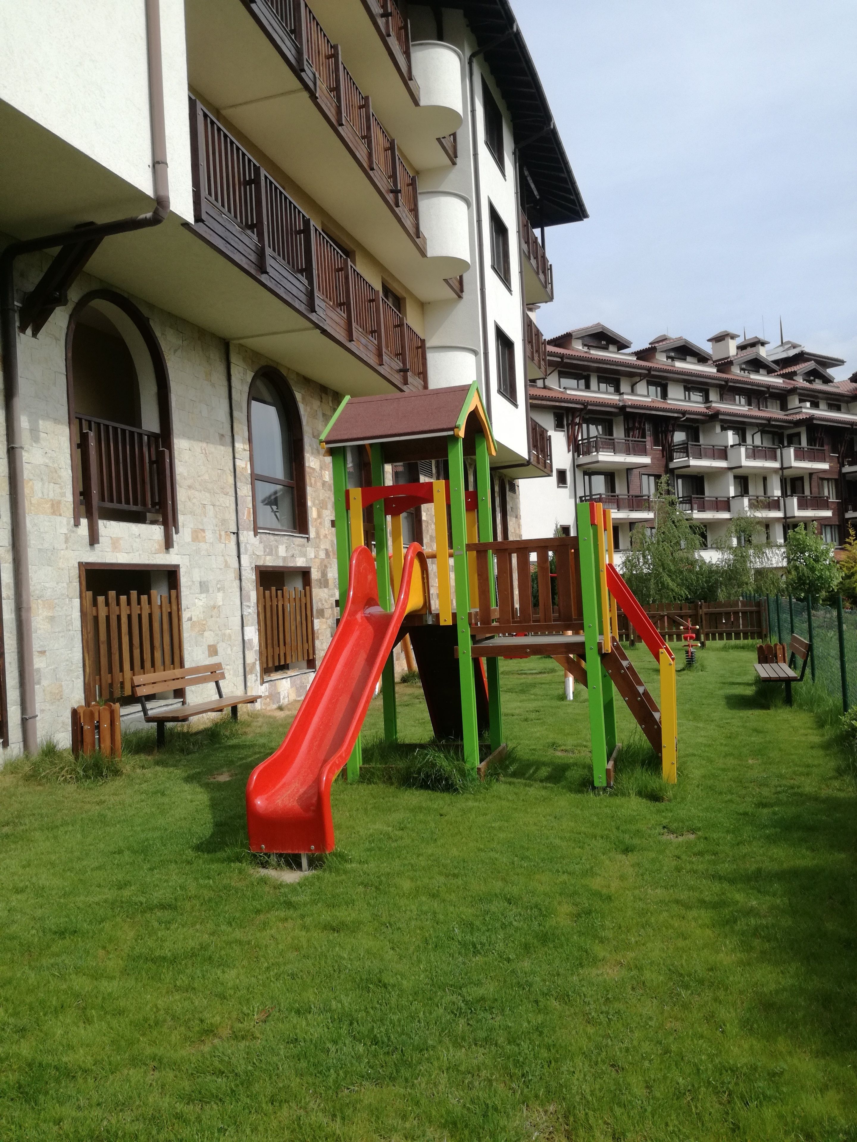 Un terrain de jeux avec un toboggan rouge et une structure d’escalade, situé devant un immeuble doté de balcons et de murs en pierre.