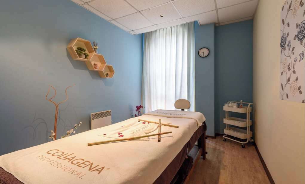 Ein Raum mit einer Massageliege, einem kleinen Regal mit Pflanzen, einer Uhr und einem Fenster mit Vorhängen.