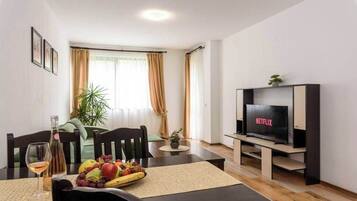 A modern living room with a dining area, a flat-screen TV, and a dining table set with fruits and a bottle of wine.
