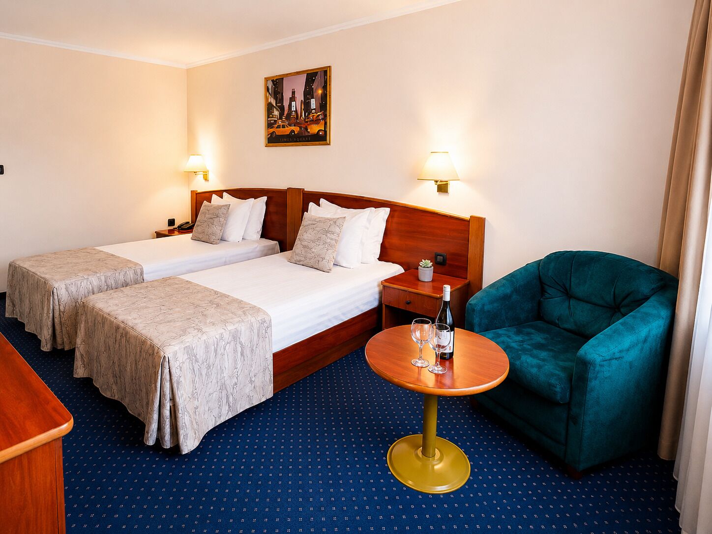 A hotel room with two beds, a small table with a bottle and glasses, a green armchair, and a painting on the wall.
