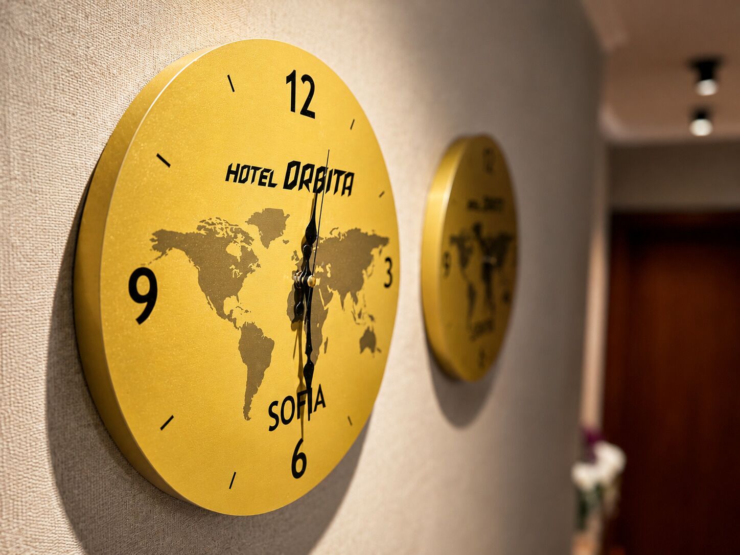 Two round wall clocks with a world map design, labeled 'HOTEL ORAITA' and 'SOFIA,' mounted on a wall.