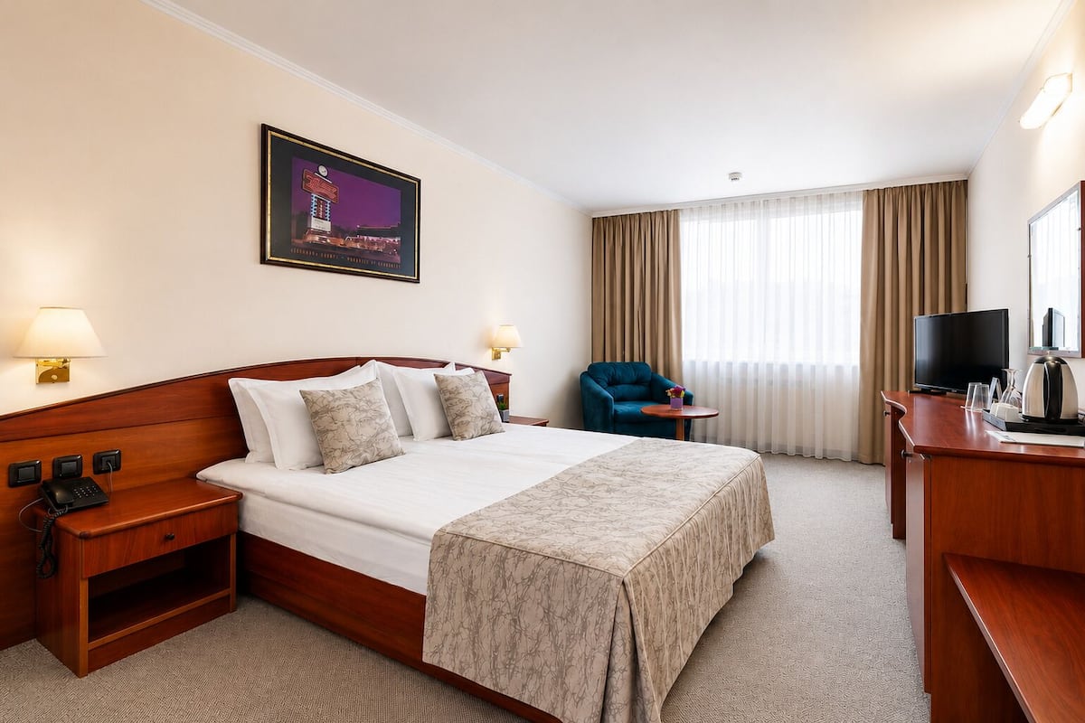 Habitación de hotel con cama, lámparas de noche, escritorio con televisor, silla y ventana con cortinas.