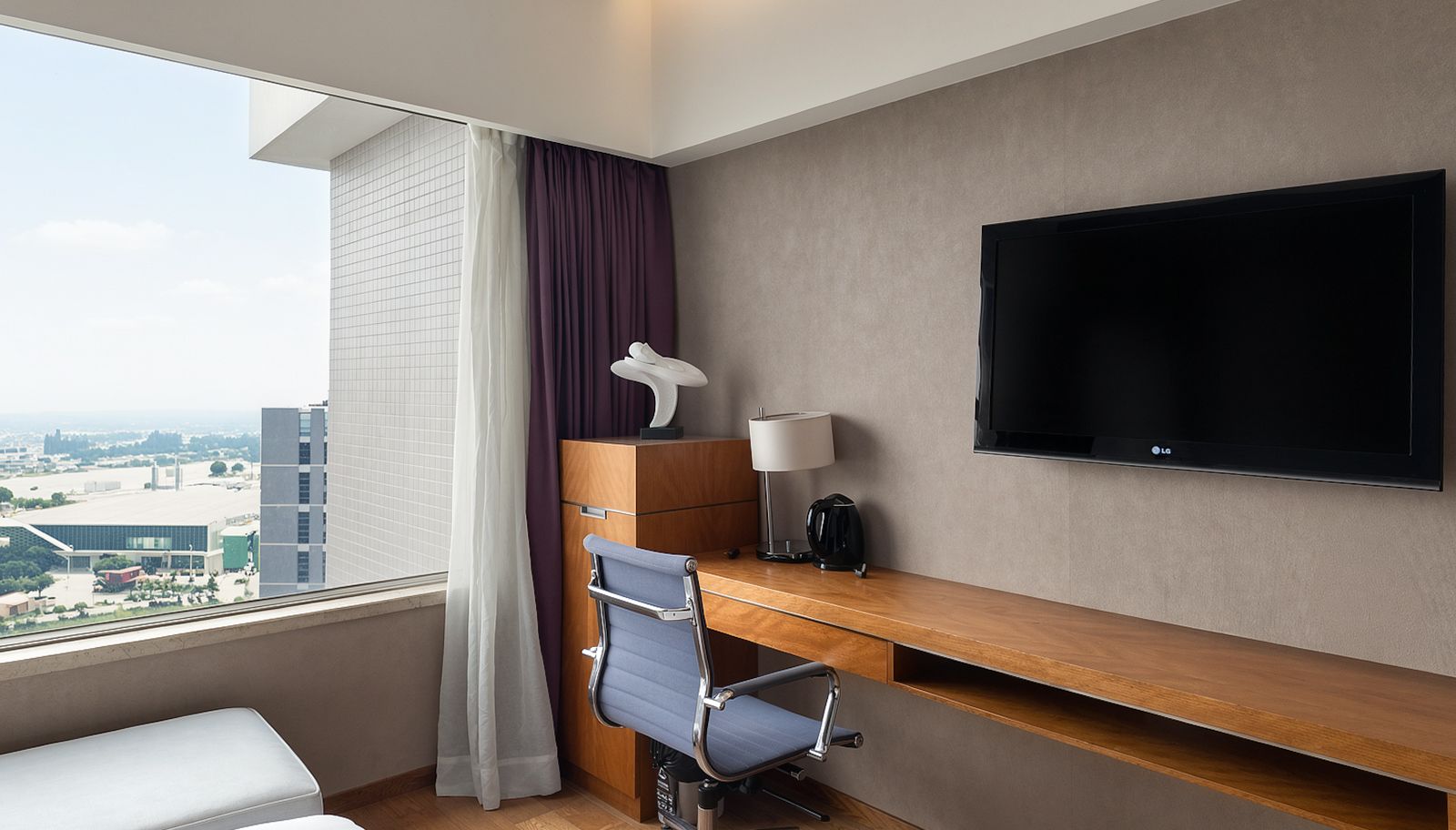 A hotel room with a desk, chair, television, and a view of the city.