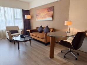 Une chambre d’hôtel moderne avec un canapé, deux fauteuils, une table basse en verre et une lampe.