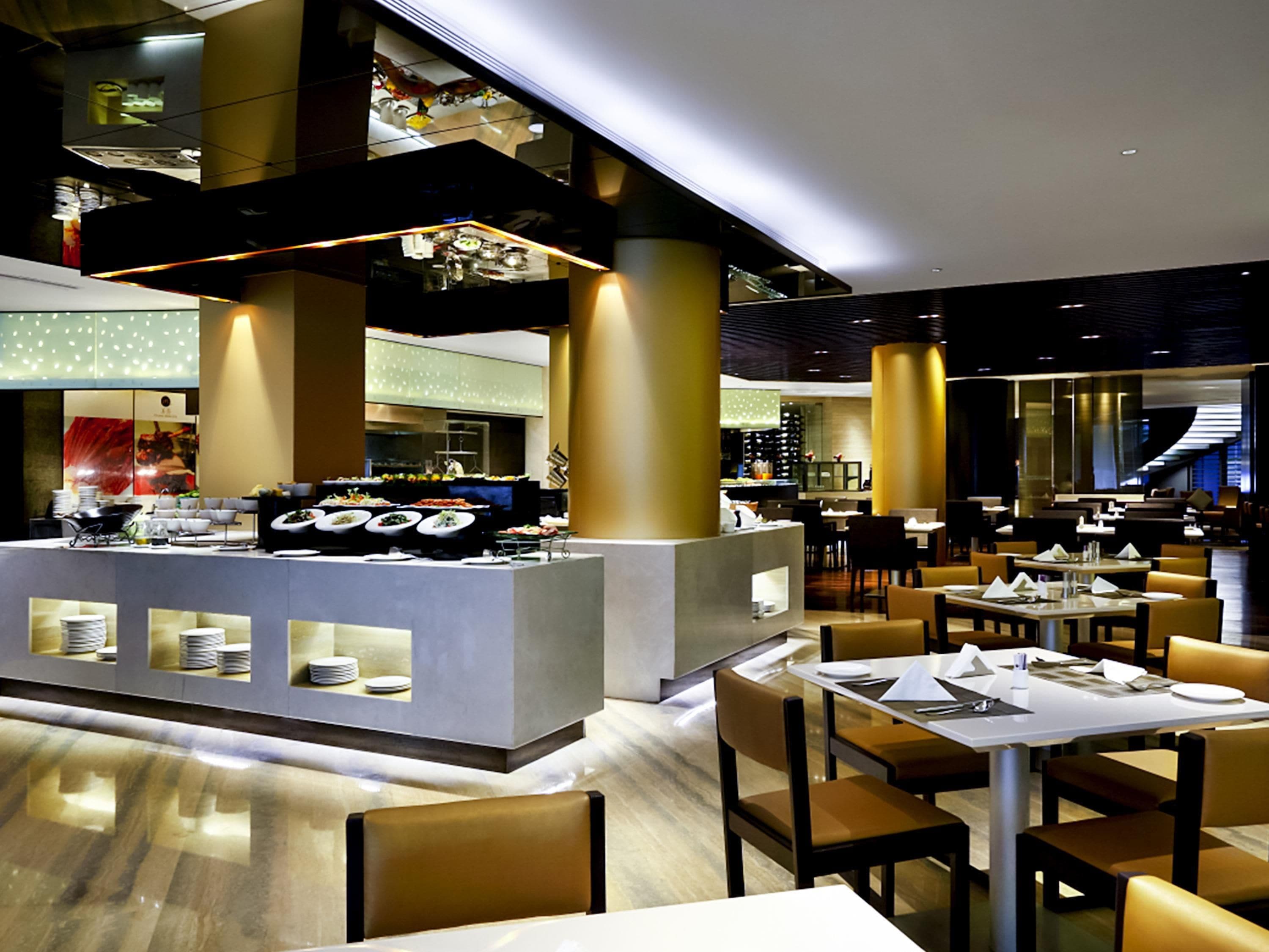 Un moderno vestíbulo de hotel con una zona de buffet, varias mesas de comedor y una escalera que lleva a un nivel superior.