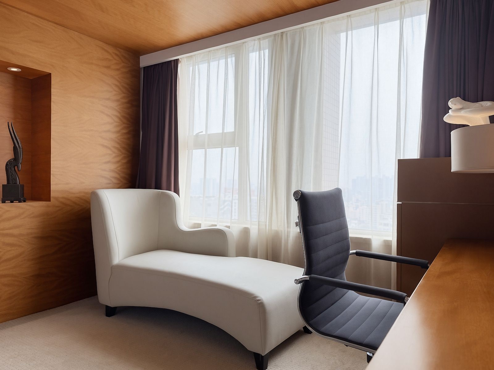 A modern hotel room with a curved white sofa, a black office chair, a wooden desk, and a large window with sheer curtains.