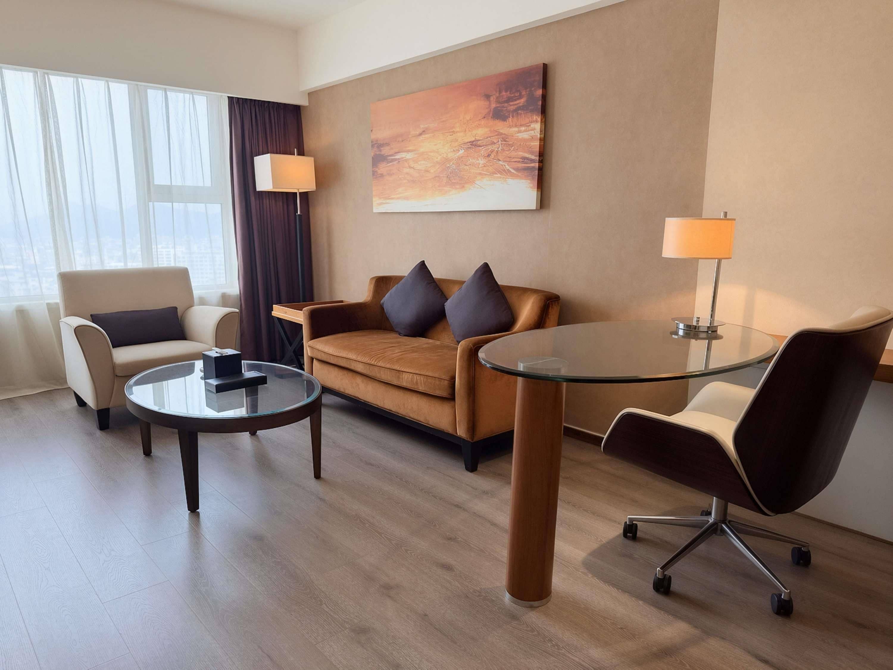 A hotel room with a sofa, armchair, and a glass-top table.