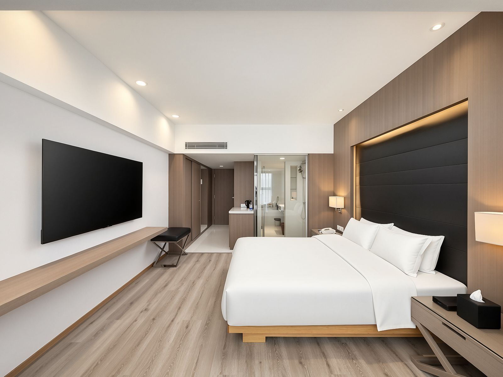 Una habitación de hotel moderna con un televisor de pantalla plana grande, una cama con ropa blanca, una mesita de noche con lámpara y un cabecero de madera.