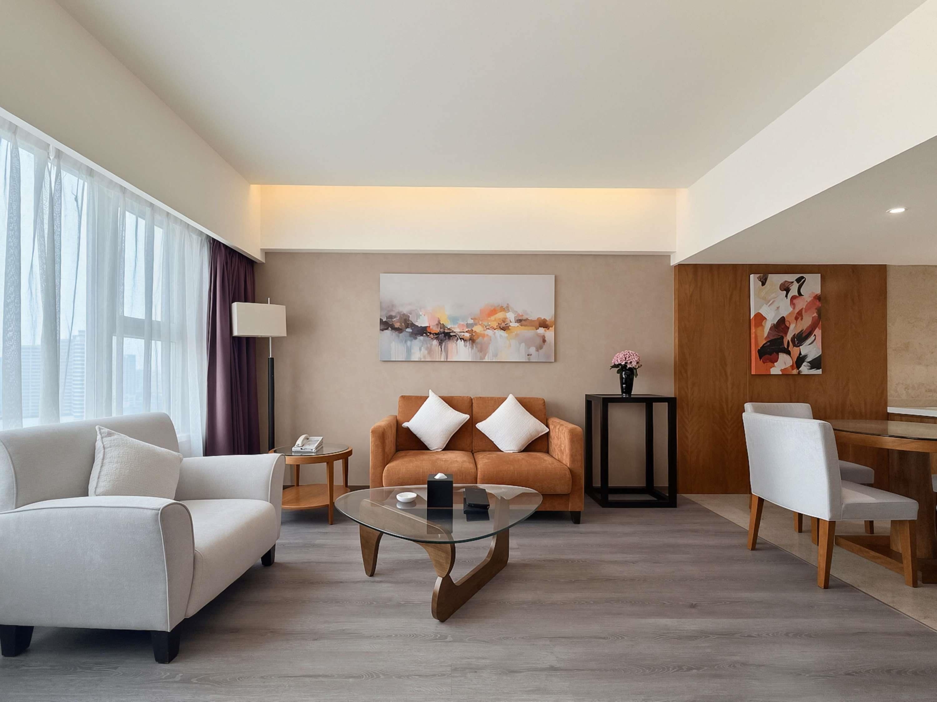 A modern hotel room with a sofa, armchair, glass coffee table, and a dining area with a chair and table.