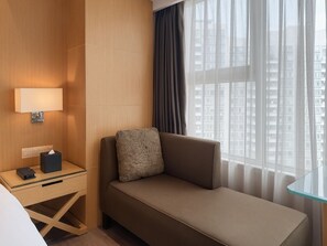 Een hotelkamer met een bank, een nachtkastje met een lamp en een uitzicht op een stadsbeeld door grote ramen.