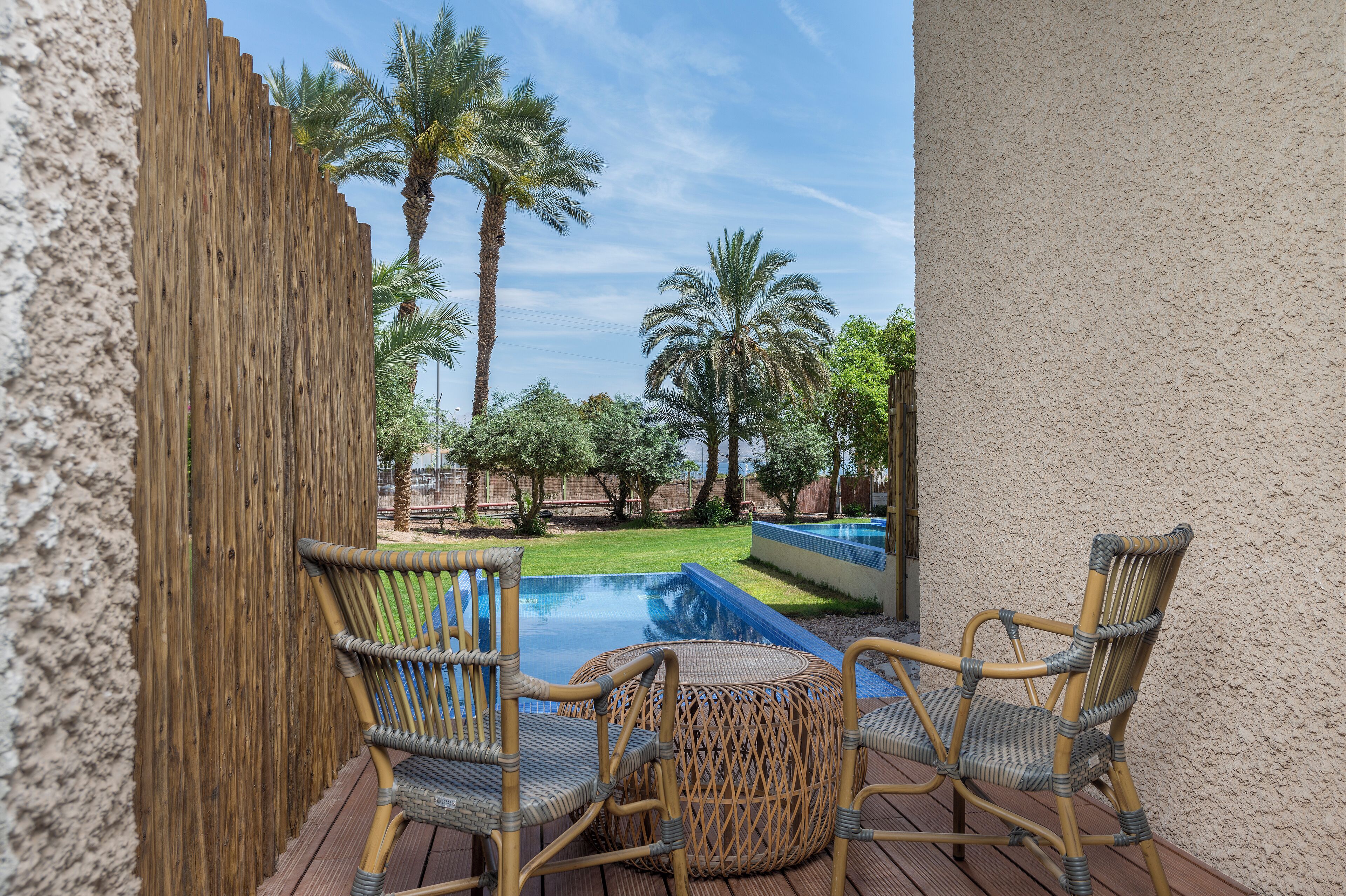 Eine Holzterrasse mit Korbstühlen und einem kleinen runden Tisch, mit Blick auf einen Swimmingpool und Palmen.