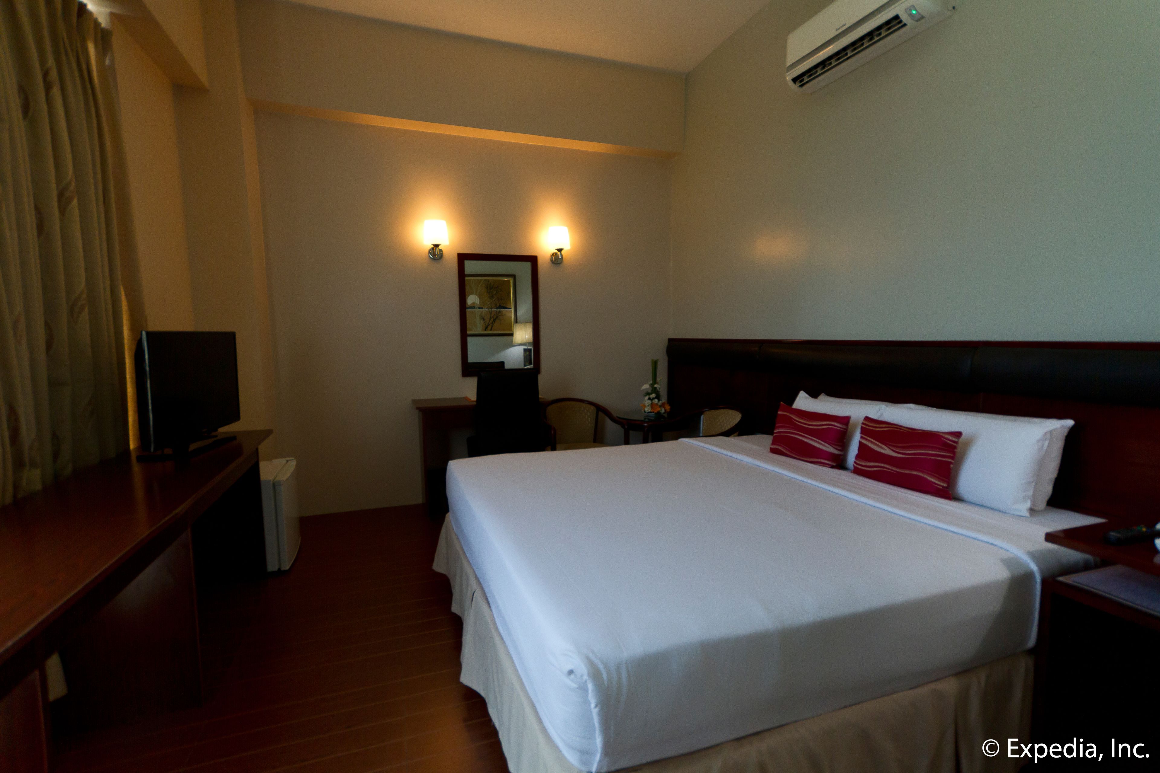 A hotel room with a large bed, a desk, a chair, a TV, and an air conditioning unit.
