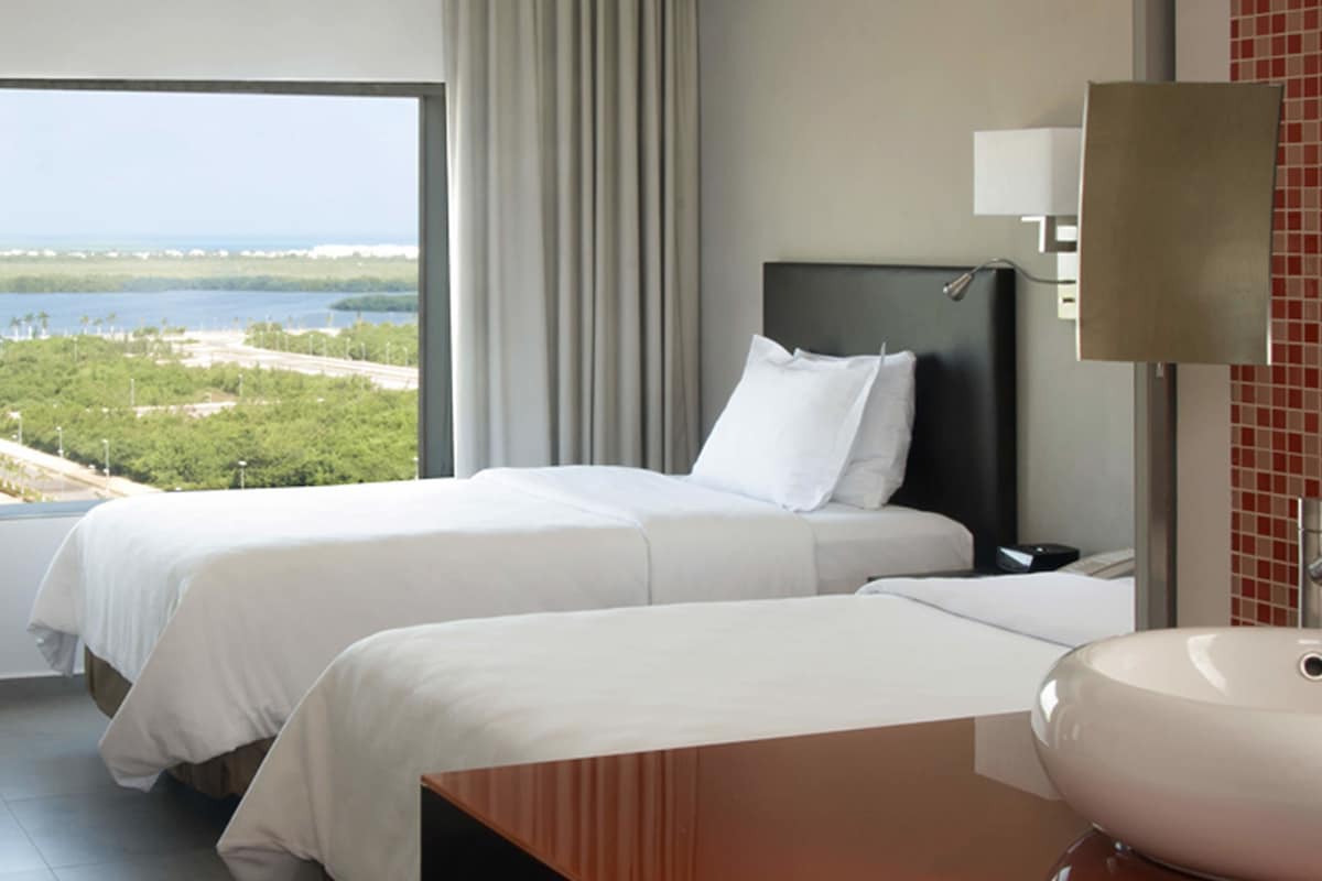 Habitación de hotel con dos camas, un ventanal con vistas al mar y vegetación, un baño con lavamanos y espejo, y una mesita de noche.