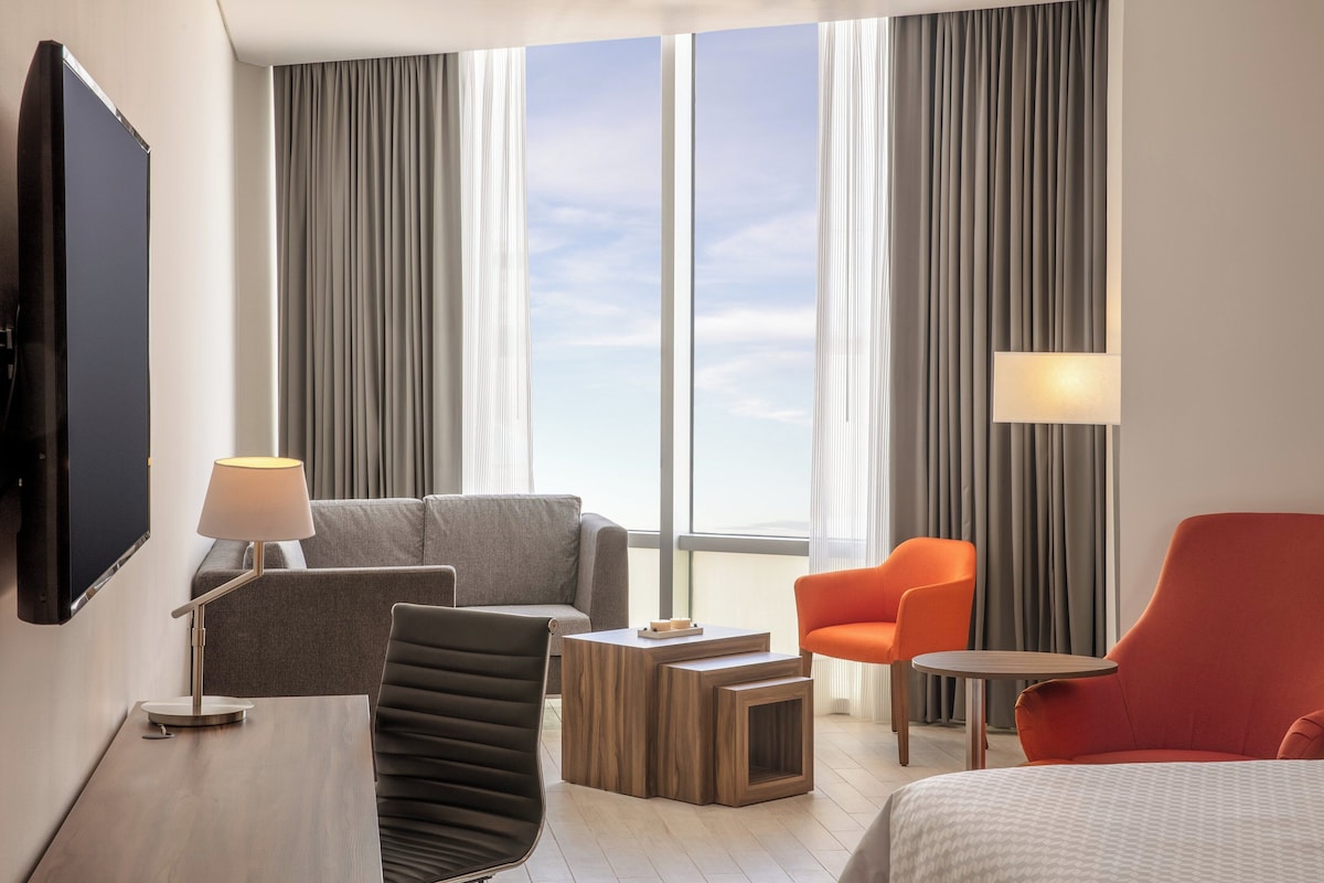 Una habitación de hotel moderna con un ventanal grande, un sofá gris, un sillón naranja, una mesa de centro de madera y un televisor montado en la pared.