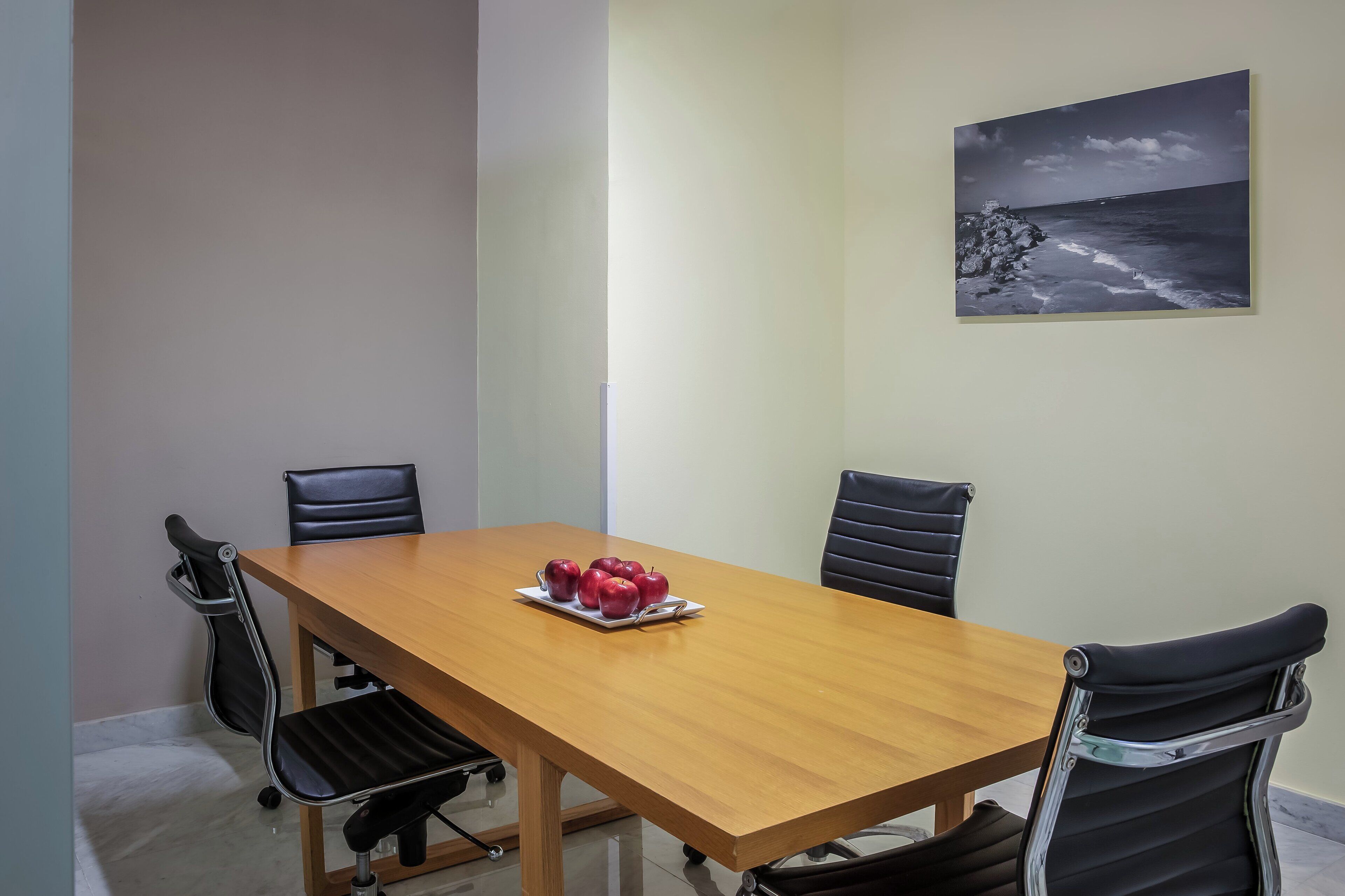 Un salón de conferencias con una mesa de madera, cuatro sillas negras y un plato de manzanas.