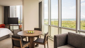 A hotel room with a large window, a round dining table with chairs, a bed, a TV, and a sofa.