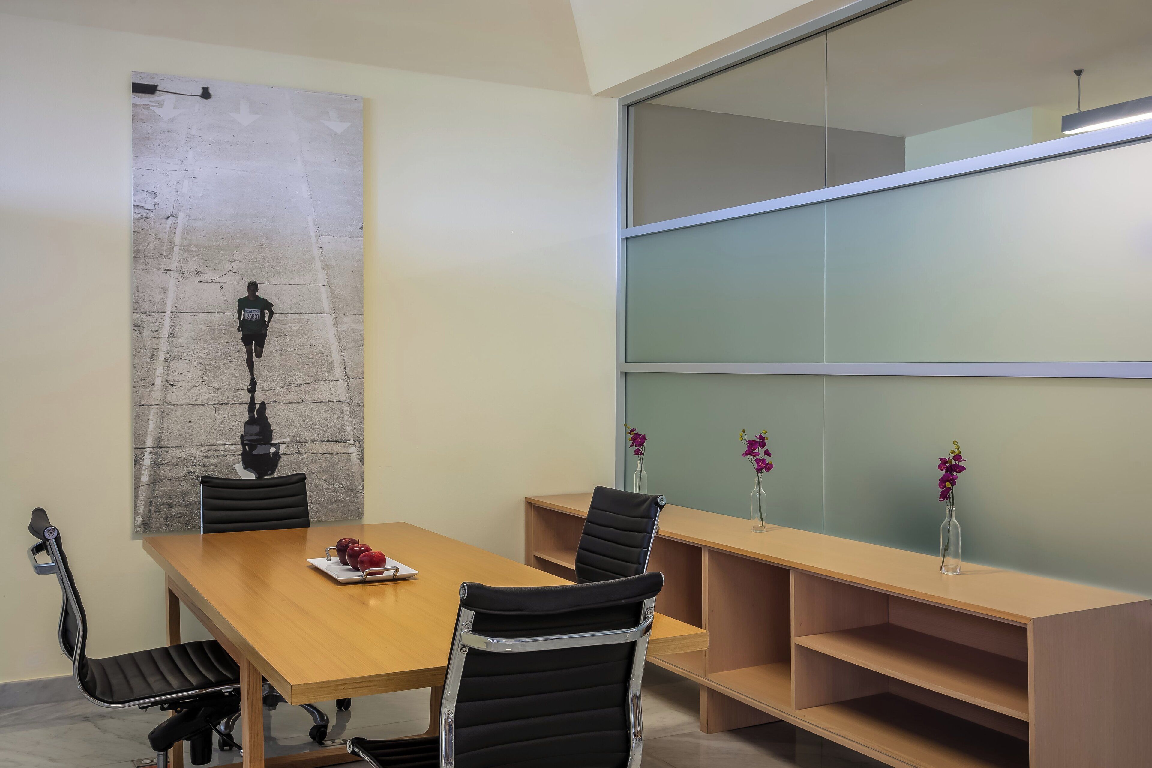 A conference room with a wooden table, black chairs, a glass partition, and a vase with flowers.