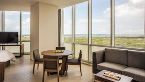 A modern hotel room with a large window, a flat-screen TV, a round dining table, and a sofa.
