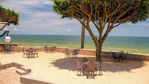 A beachside dining area with tables and chairs, a tree, and a view of the sea. - Protea Hotel by Marriott Entebbe (Entebbe)