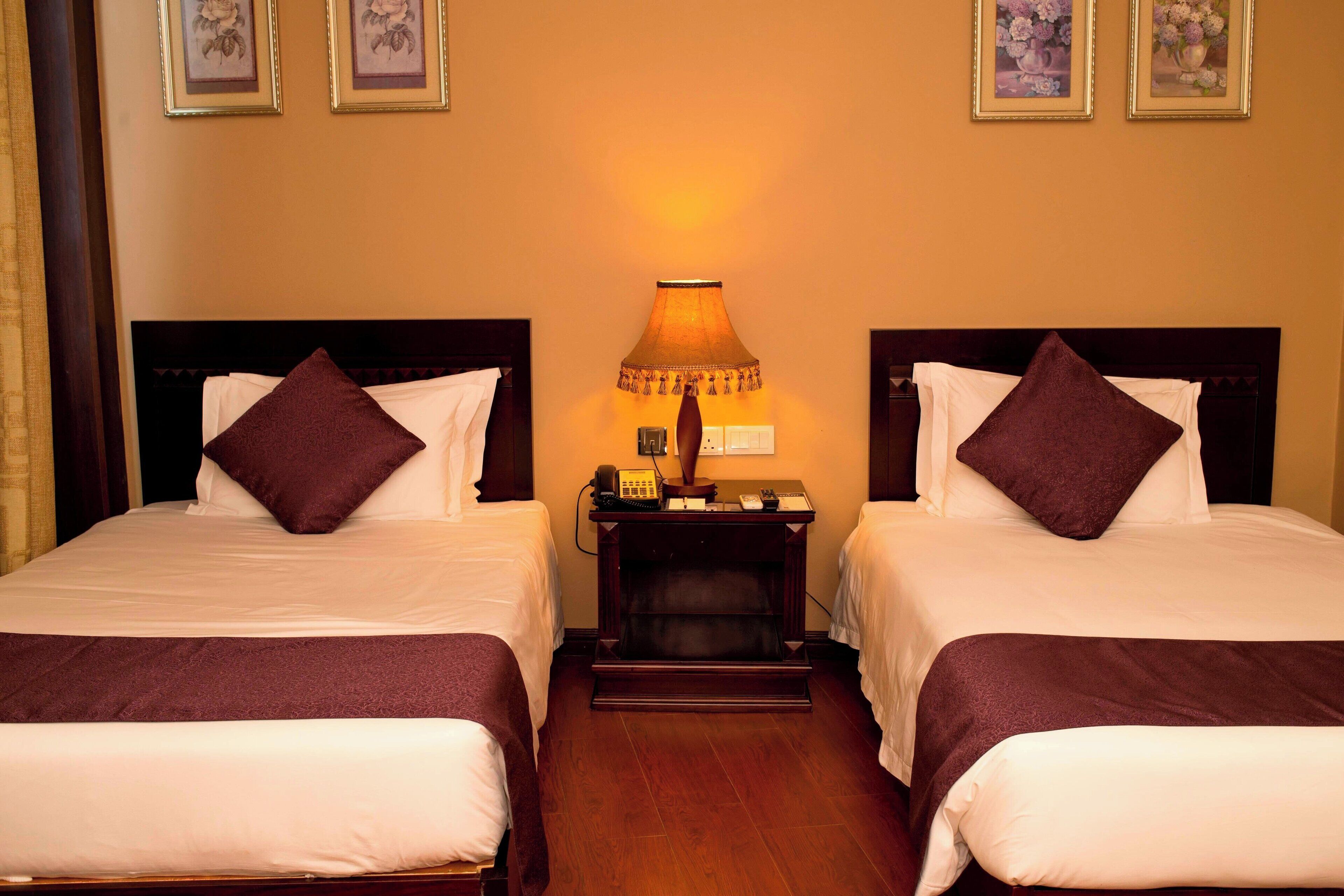 A hotel room with two beds, each with a dark brown headboard and pillow, a bedside table with a lamp, and framed floral artwork on the wall.