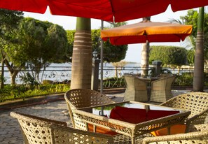 Wicker chairs and a table under a red umbrella, with a view of a body of water and greenery in the background. - Protea Hotel by Marriott Entebbe (Entebbe)