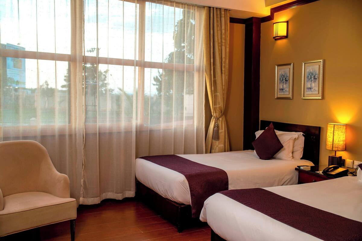 Una habitación de hotel con dos camas, una silla, una mesita de noche con lámpara y una ventana con cortinas.
