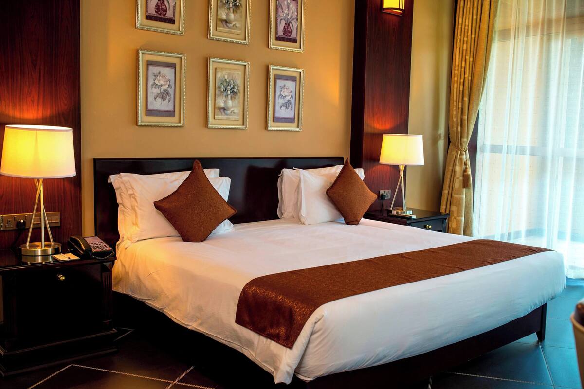 Habitación de hotel con una cama grande, dos mesitas de noche, lámparas, cuadros en la pared y un ventanal con cortinas.
