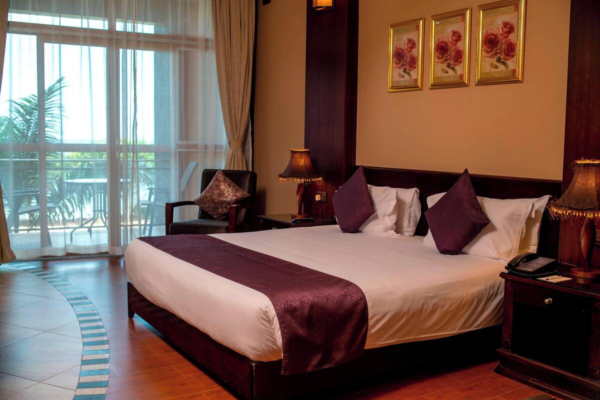 Habitación de hotel con una cama grande, dos mesitas de noche, una silla y vistas al exterior.