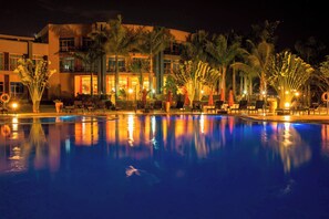 Vista nocturna de una zona de piscina de hotel, con el reflejo del edificio y palmeras.