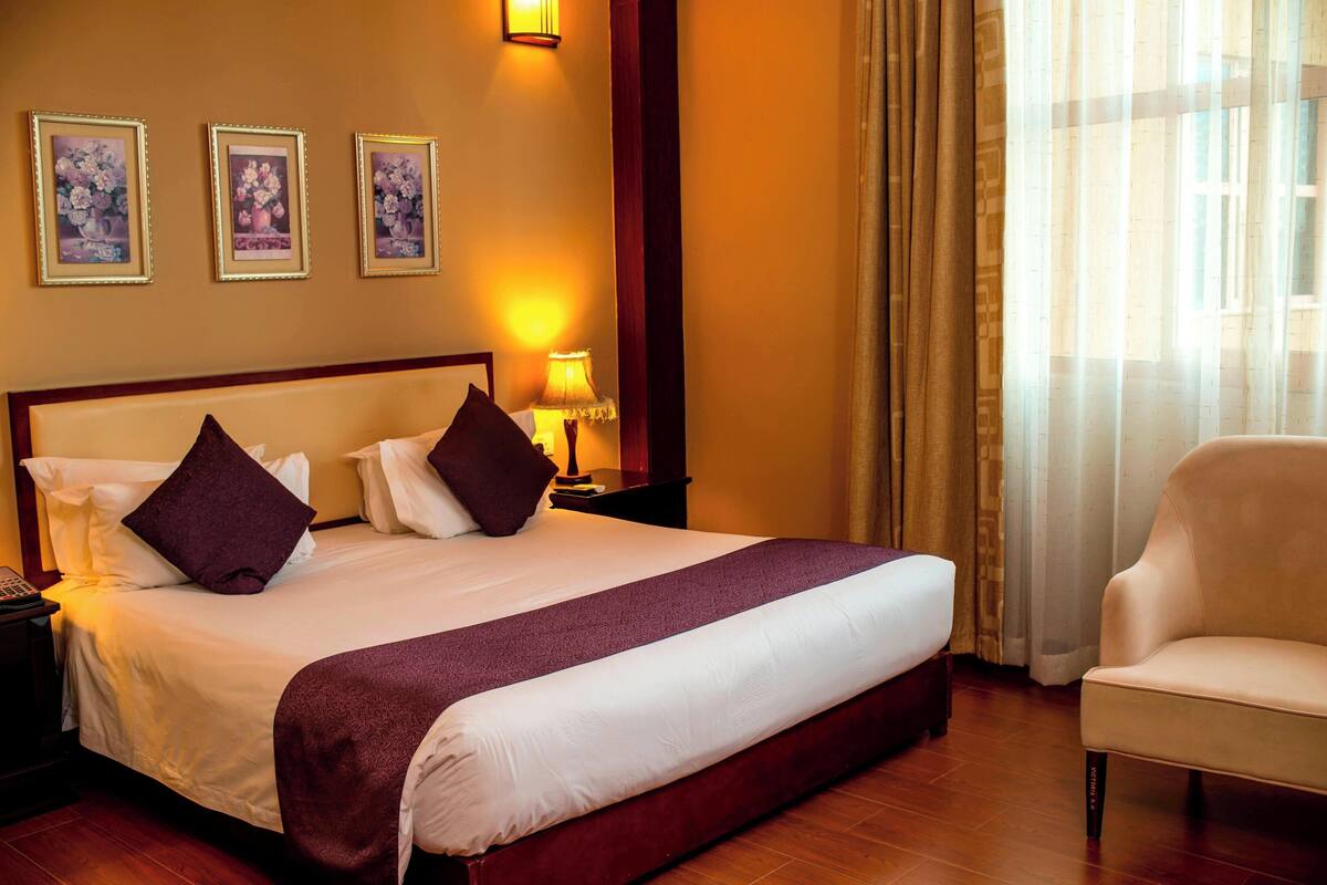 Habitación de hotel con una cama grande, dos almohadas, una mesita de noche con lámpara, una silla y tres cuadros en la pared.