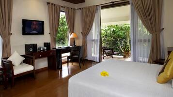 A hotel room with a bed, desk, chair, TV, and a balcony with plants.
