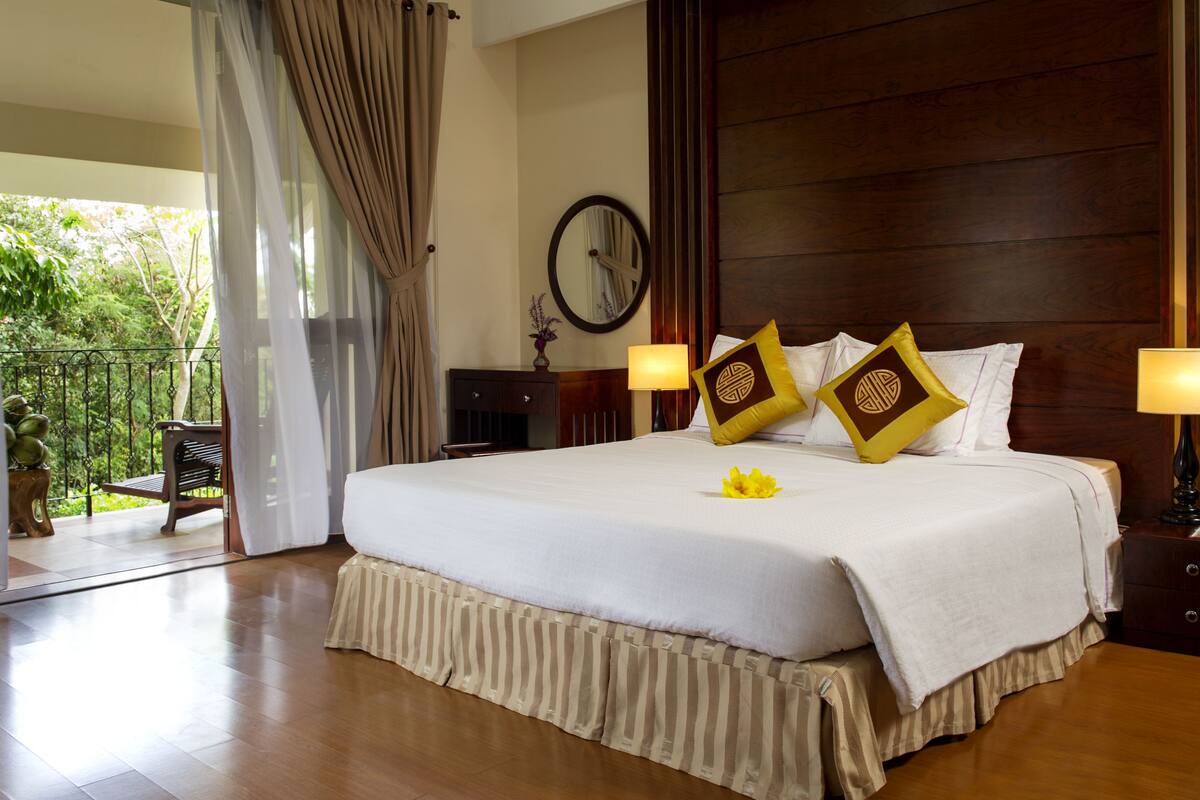 A large bed with white linens and two decorative pillows, a bedside table with a lamp, a wooden headboard, a round mirror, and a view of greenery through a glass door.